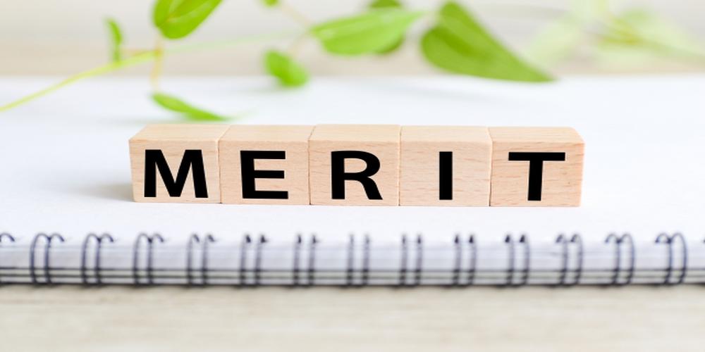 木製ブロックに「MERIT」の文字が並び、背景にノートとグリーンが映る明るい構成