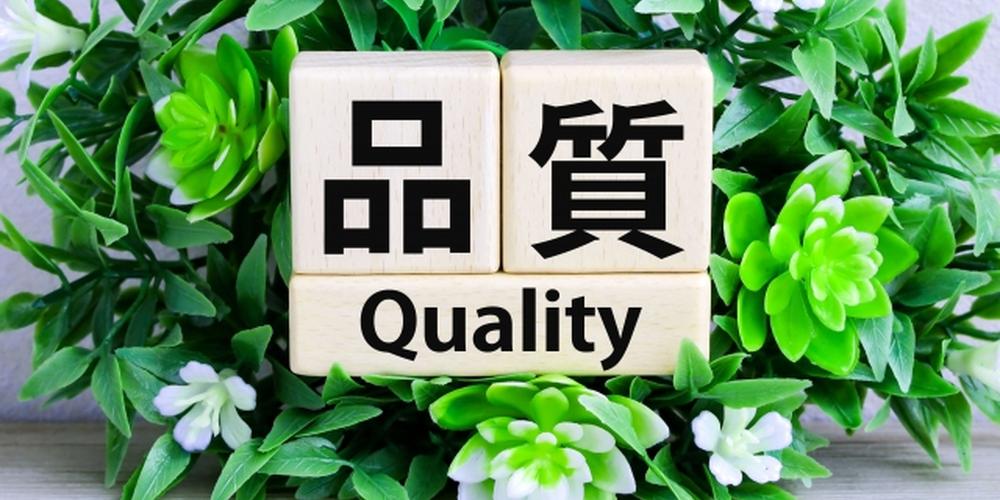 木製ブロックに「品質」と「Quality」と書かれた文字と緑の植物が背景にあるイメージ