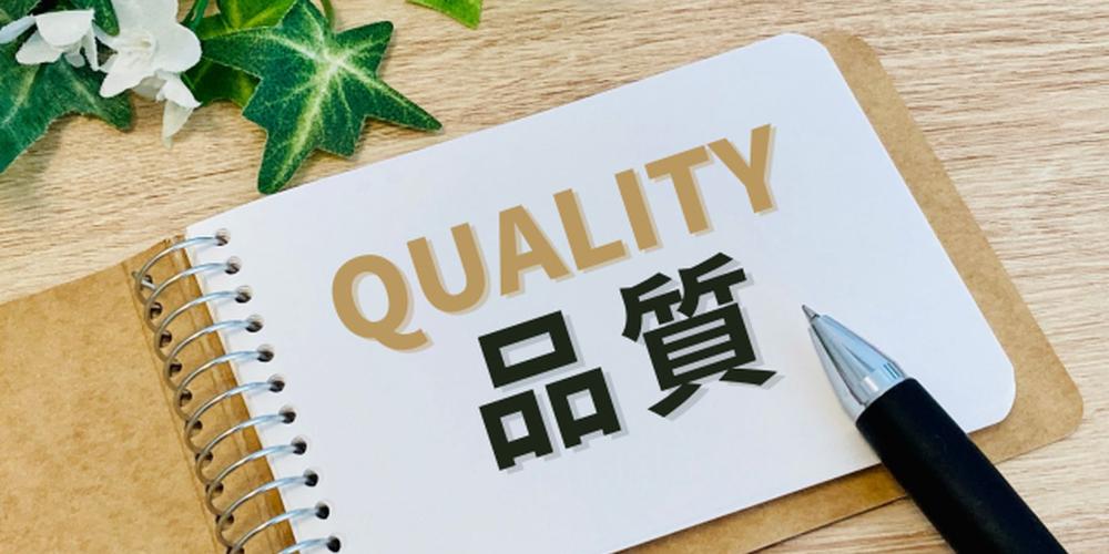 ノートに書かれた「QUALITY 品質」の文字と黒いボールペン、観葉植物が並ぶ高品質イメージのデスク写真