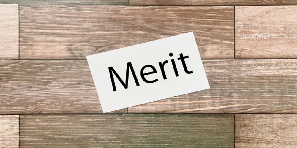 木目調の背景に「Merit」と書かれた白いカードが斜めに置かれている画像（メリットのイメージ）