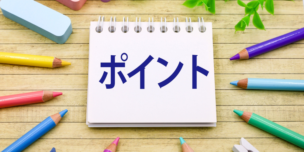 カラフルな色鉛筆が周囲に散りばめられたデスクの上に、ノートに青文字で「ポイント」と書かれているシンプルな写真