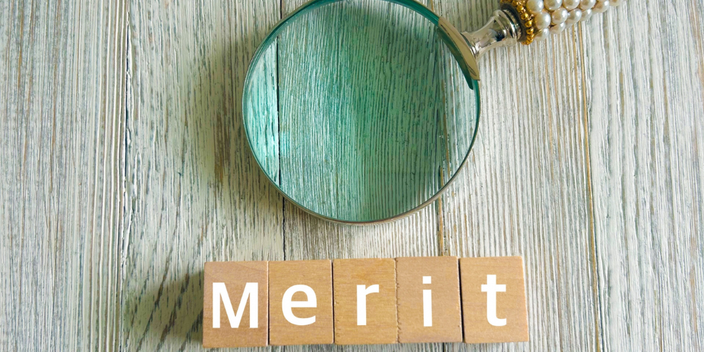 木目の机上に虫眼鏡が伏せられ、その下に木製ブロックで白抜き文字の「Merit」が並ぶ写真