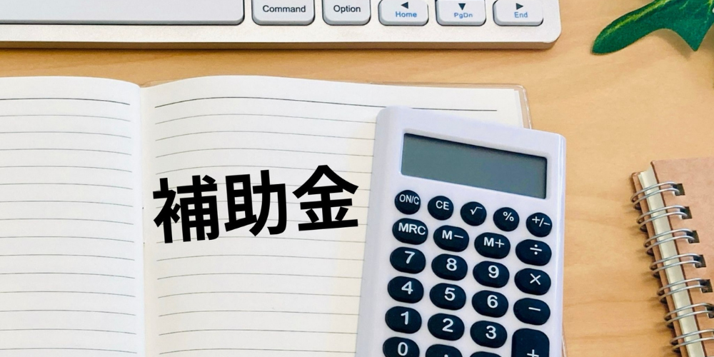 ノートを開いた机上に電卓とリングノート、パソコンのキーボードが置かれ、ノート面に「補助金」という文字が重ねられた事務作業イメージ画像
