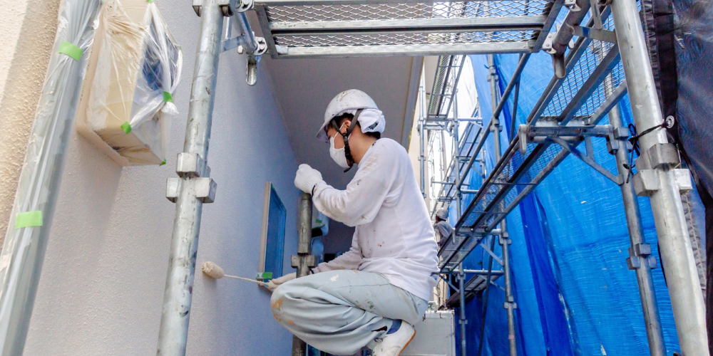 足場上で白い作業着とヘルメットを着用した塗装職人が壁をローラーで塗装している作業風景