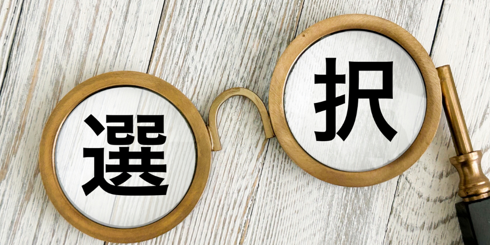 木製の丸メガネのレンズ部分に「選」と「択」という文字がそれぞれ映り込み、木目のテーブル上にメガネ型拡大鏡が置かれた選択を示すイメージ画像