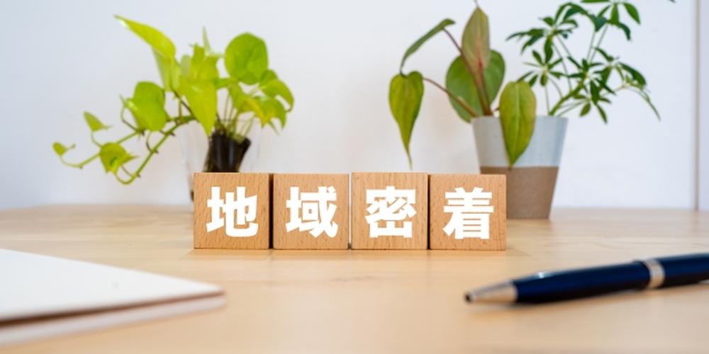 観葉植物が並ぶ机の上に「地域密着」と書かれた木製ブロックが置かれているオフィスイメージ